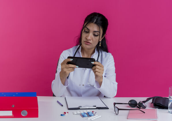 年轻人令人印象深刻的年轻女医生穿着医用长袍和听诊器坐在办公桌旁 手持医疗工具 看着隔离在粉红色墙上的手机工具长袍医疗
