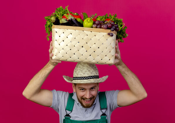 帽子快乐的年轻英俊的斯拉夫园丁 穿着制服 头上戴着帽子 手里拿着一篮蔬菜 被隔离在深红色的墙上年轻蔬菜深红色