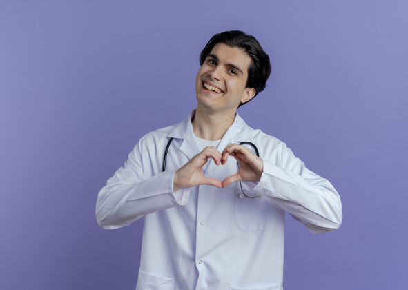 心脏快乐的年轻男医生穿着医用长袍和听诊器在紫色的墙上做着隔离的心脏标志 还有复制空间听诊器穿着医学