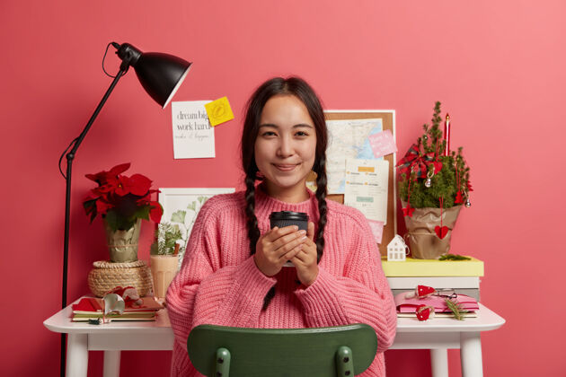 桌子迷人的韩国女孩拿着外卖咖啡 坐在办公室附近的椅子上 完成工作 贴在粉红色的墙上女人喝酒毛衣