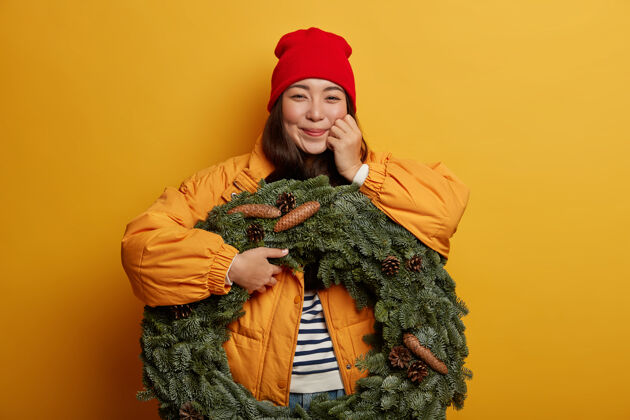 卷发快乐的韩国女人穿着冬天的外套 表达真诚的情感 手持美丽的云杉花环 站在室内黄色的背景下自然举行室内