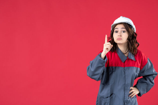 制服穿着制服 戴着安全帽 指着孤立的红墙 惊讶的女建筑工人的特写镜头硬女士尖