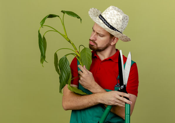 英俊困惑的年轻英俊的斯拉夫园丁穿着制服 拿着帽子 看着橄榄绿墙上孤立的植物花园斯拉夫帽子