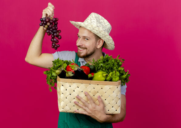 拿着面带微笑的年轻英俊的斯拉夫园丁 身着制服 戴着帽子 手里拿着一篮子蔬菜和葡萄 隔离在深红色的墙上 留着复制空间年轻深红色看着