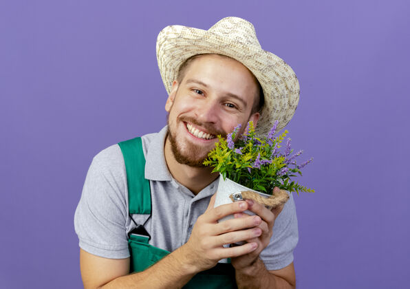 帽子微笑着的年轻英俊的斯拉夫园丁 身着制服 戴着帽子 手持花盆 在紫色的墙壁上 看起来与世隔绝 有复制空间花园斯拉夫人花盆
