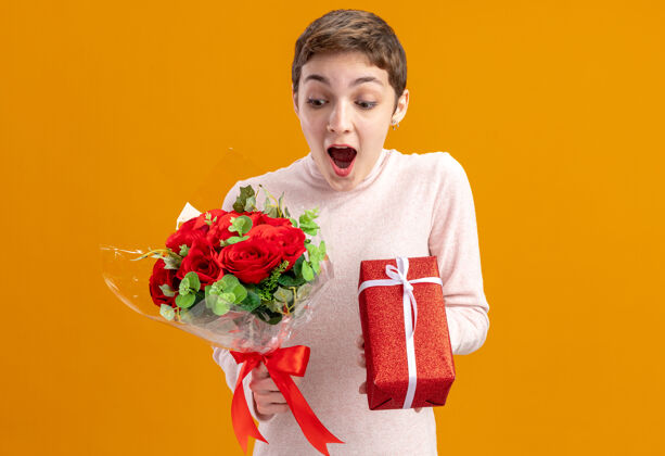 概念一位留着短发的年轻女子手持一束红玫瑰 手里拿着一件礼物 站在橘色的墙上 看上去既惊讶又惊讶女人年轻情人节