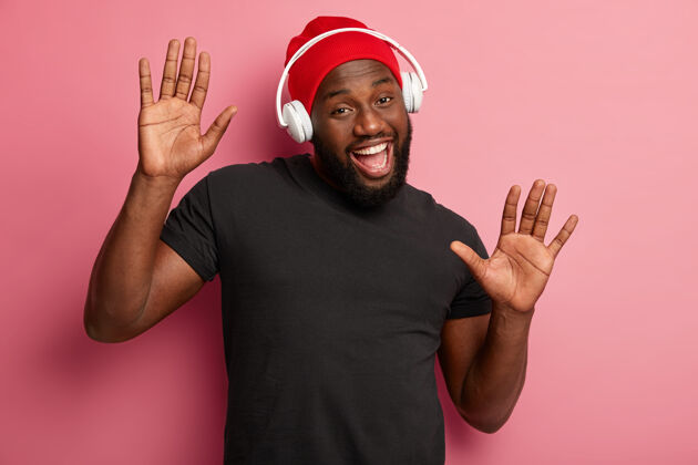 快乐满意的胡须男青年戴着耳机听欢快的歌曲 在粉色背景上移动 用酷音乐提升心情 感觉乐观 戴着红帽子和黑色t恤嘴肖像休闲