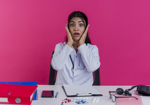 长袍震惊的年轻女医生穿着医用长袍和听诊器坐在办公桌旁 手放在隔离在粉红色墙上的脸上工具医生听诊器