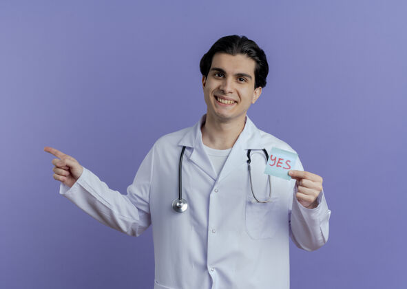 穿面带微笑的年轻男医生穿着医用长袍 听诊器显示“是”的字条 指向紫色墙壁上隔离的一侧 并留有复印空间听诊器医生展示