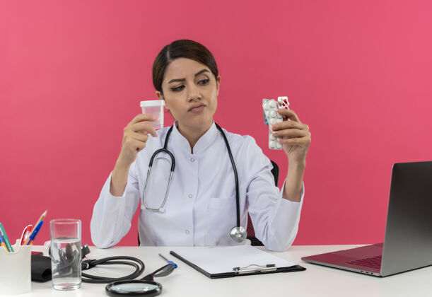 烧杯可疑的年轻女医生穿着医用长袍和听诊器坐在办公桌旁 手里拿着医疗工具和笔记本电脑 看着隔离在粉红色墙上的药品和烧杯年轻拿着药物