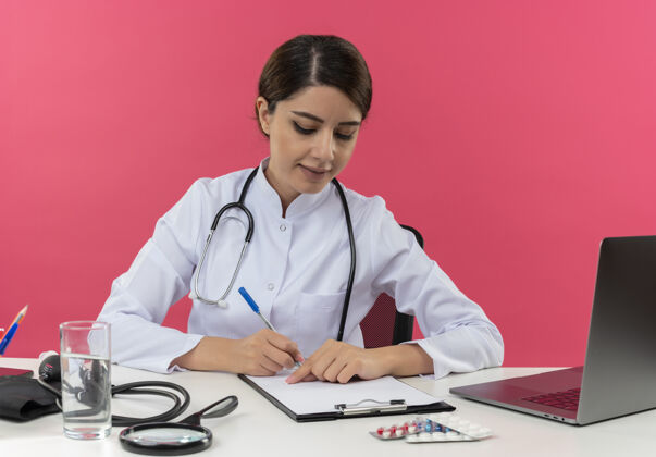 坐着专注的年轻女医生穿着医用长袍和听诊器坐在办公桌旁 手里拿着医疗工具和笔记本电脑 写字板上的笔孤立地写在粉红色的墙上听诊器医疗年轻人