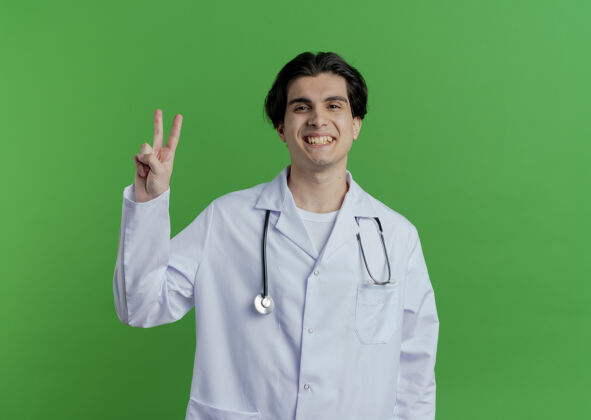 男性微笑着的年轻男医生穿着医用长袍和听诊器做和平的标志隔离在绿色的墙壁与复制空间穿微笑和平