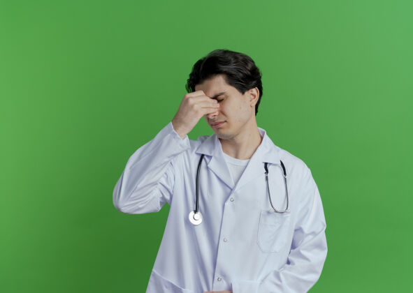 持有疲惫不堪的年轻男医生穿着医用长袍 手持听诊器 闭着眼睛闭着鼻子 隔离在绿墙上 留着复印空间男性鼻子穿