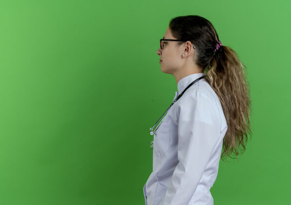 查看年轻的女医生穿着医用长袍 戴着听诊器和眼镜站在侧面图上 看着笔直的隔离在绿色的墙上 有复印空间女眼镜个人资料