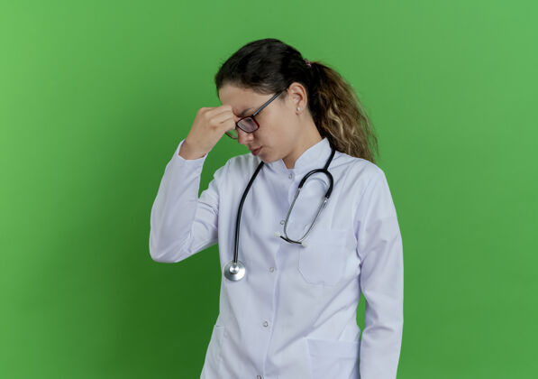 持有疲惫的年轻女医生穿着医用长袍 戴着听诊器 戴着眼镜 闭着眼睛抱着鼻子 隔离在绿墙上 留着复印空间医生长袍疲倦