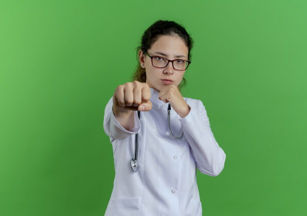 自信自信的年轻女医生穿着医用长袍 戴着听诊器 戴着眼镜 在绿色的墙上做着拳击动作 并留有复制空间医生长袍穿着