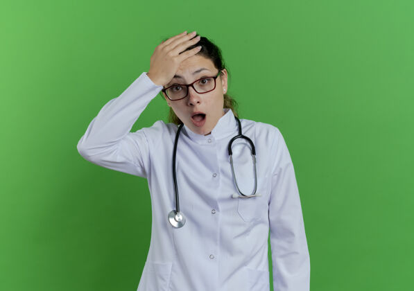 手年轻的女医生穿着医用长袍 戴着听诊器 戴着眼镜 手放在头上 被隔离在绿色的墙上 留着复印空间医学惊讶听诊器