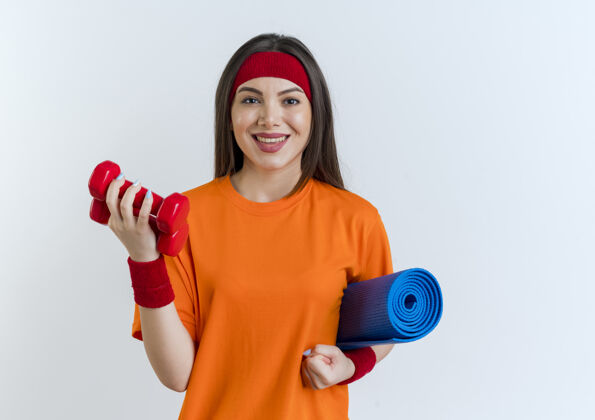 哑铃面带微笑的年轻运动女性戴着头带和腕带拿着瑜伽垫和哑铃隔离在白色的墙壁上 有复制空间运动垫子腕带