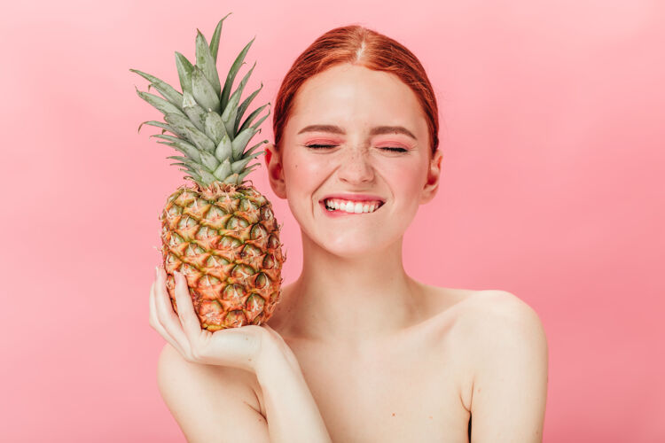 微笑正面图是一个快乐的女人 她闭着眼睛摆着菠萝的姿势摄影棚拍摄的是一个兴奋的姜汁女孩拿着粉红色背景上的水果年轻粉色粉色背景