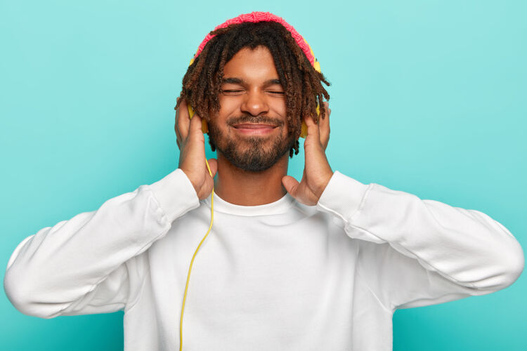 轨道时尚的男人专注于歌曲 双手放在现代耳机上 享受响亮的声音 闭上眼睛远离快乐 穿着白色休闲套头衫娱乐室内人