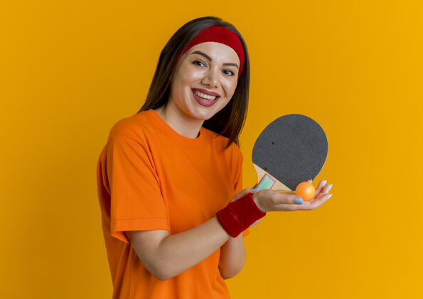 年轻快乐的年轻运动女性戴着头带和腕带拿着乒乓球拍和球看握球拍女子