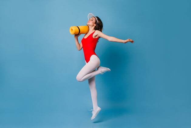 头部穿着红色紧身衣和白色连裤袜的苗条黑发女士手持瑜伽垫80后活动移动