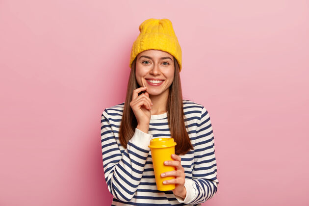 休闲可爱的女人喜欢喝咖啡 拿着外卖杯 愉快地微笑 戴着黄色的帽子和条纹毛衣 在粉红色的墙上摆姿势 享受气味墙壁时尚肖像