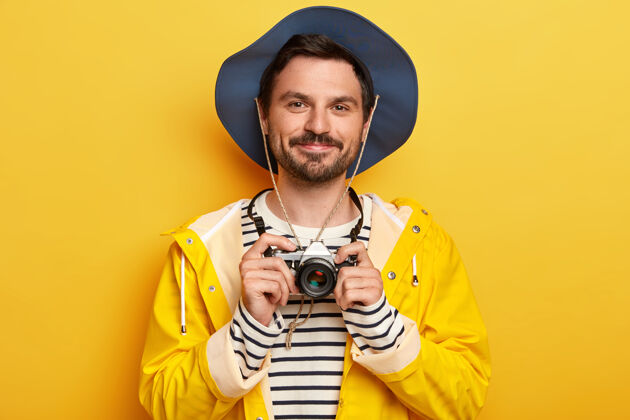 肖像帅气的没刮胡子男人的肖像拿着复古相机 拍照 戴着帽子 条纹套头衫和雨衣 隔离在黄色的墙上胡子雨衣持有