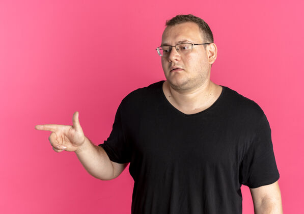 手指戴眼镜的超重男子身穿黑色t恤 表情严肃 手指指向粉色上方的一侧T恤壁板靠边