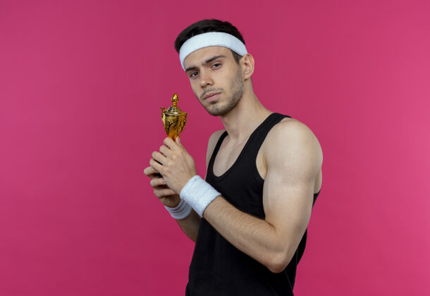 表情戴着头巾的年轻运动型男子拿着奖杯 表情自信严肃地站在粉红色的墙上奖杯运动男人