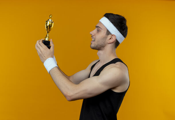 积极戴着头巾的年轻人拿着他的奖杯 站在橘色的墙上 面带微笑地看着我男人年轻运动