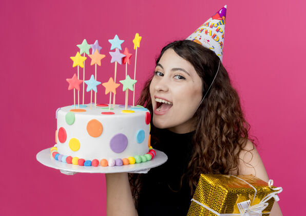 兴奋一位年轻漂亮的女士 卷发 戴着节日帽 手里拿着生日蛋糕和礼盒 生日派对的概念是粉色的女人盒子年轻