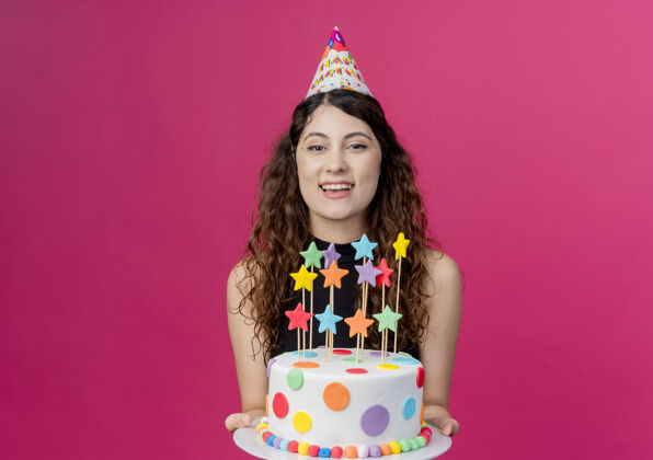 女人一位年轻漂亮的女士 一头卷发 戴着节日帽 手里拿着生日蛋糕 幸福快乐的生日派对概念在粉色之上幸福看站着
