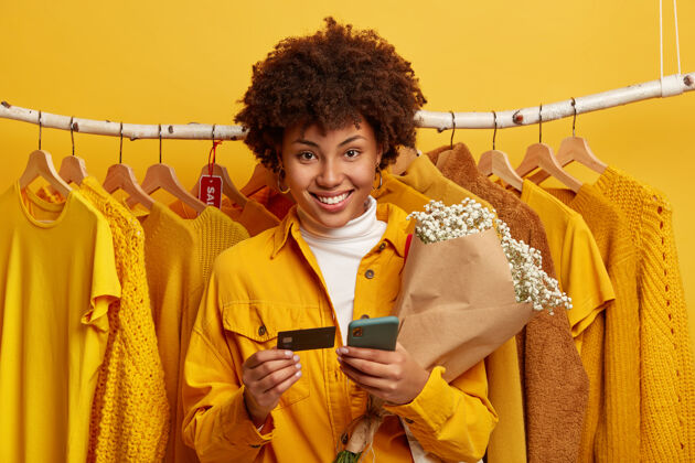 年轻快乐的非洲女人拿着手机 信用卡和花束 站在衣架上服装电子沟通