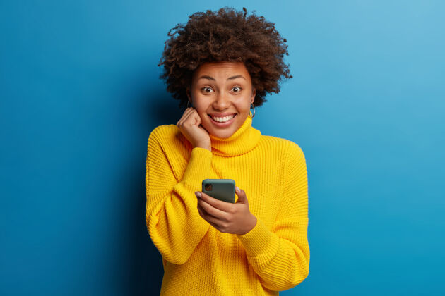 编织可爱的黑皮肤成年女子穿着黄色套头衫用手机带着幸福的表情通讯短信水平
