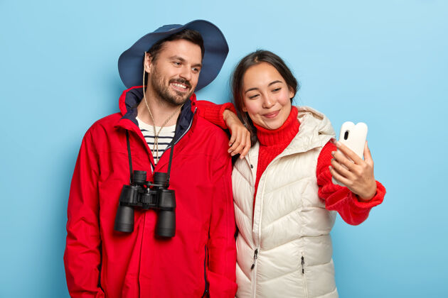 电话快乐的混血可爱情侣 表情愉快 用智能手机相机自拍 一起度假 穿着随意 用望远镜帽子背心女性