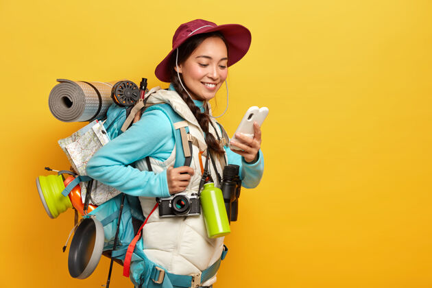 科技积极漂亮的亚洲游客使用现代小玩意导航 戴着帽子和夹克衫 使用望远镜 复古相机 卡雷马特在旅行中站在黄色的墙壁地图休息女人