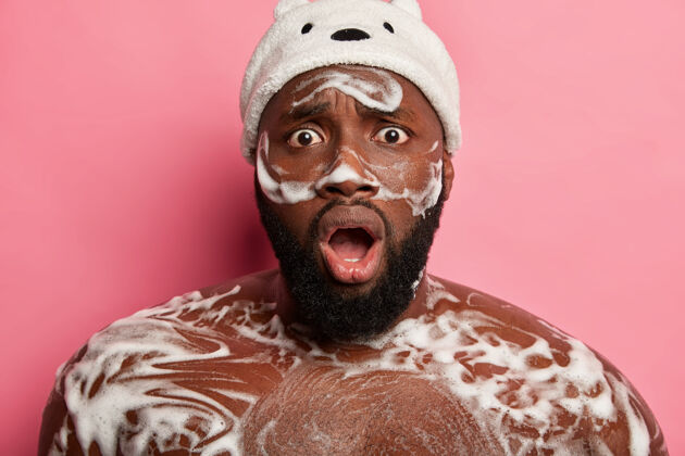 身体尴尬的深色皮肤的家伙在淋浴时洗身 皮肤上有泡沫 被粉色背景下令人惊异的东西惊呆了震惊胡须室内