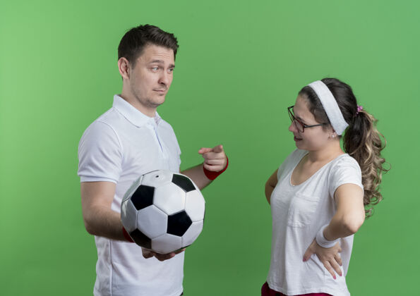 足球年轻的运动情侣严肃的男子拿着足球看着他微笑的女友在绿色微笑球男人