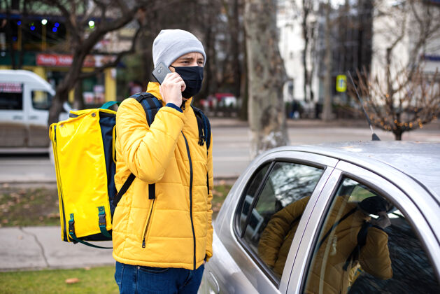 感染带着黄色背包和黑色医用面罩的快递员在车旁打电话快递街道在线