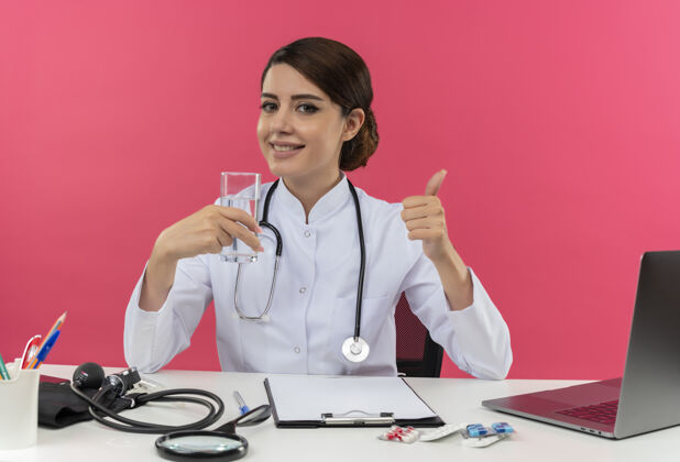 拿着面带微笑的年轻女医生穿着医用长袍和听诊器坐在办公桌旁 手里拿着医疗工具和笔记本电脑 手里拿着一杯水 竖起大拇指专业听诊器女