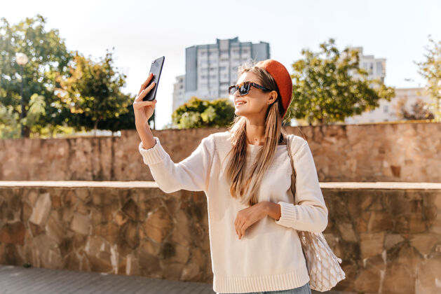 时尚穿着白衬衫的法国女士在秋天的周末自拍穿着红色贝雷帽的可爱女孩拿着手机站在街上法国贝雷帽女性