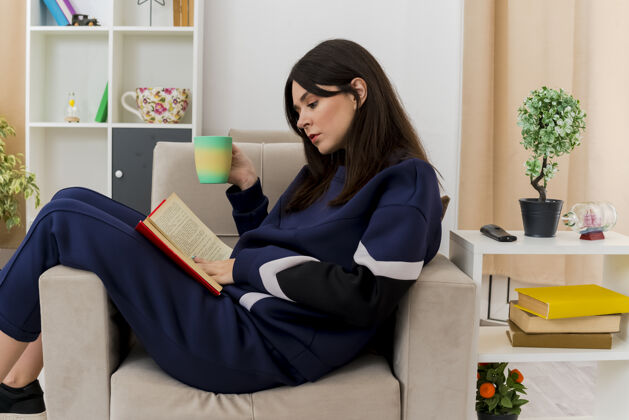 触摸年轻漂亮的白人妇女坐在设计客厅的扶手椅上 手里拿着杯子 腿上放着书 抚摸着书抱着杯子坐着