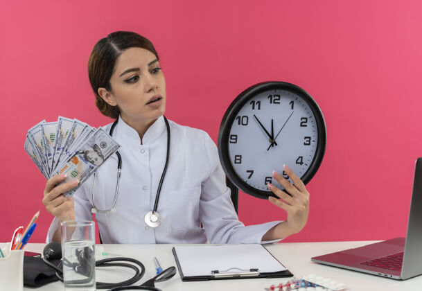 办公桌印象深刻的年轻女医生穿着医用长袍 听诊器坐在办公桌旁 手里拿着医疗工具和笔记本电脑 手里拿着钱和时钟看着时钟女人笔记本电脑制服