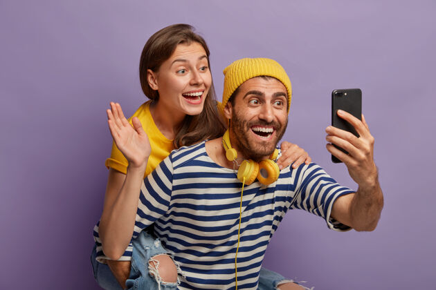 女士快乐的小两口打视频电话 面前拿着智能手机 小伙子背着在手机摄像头里挥手的女友 在紫色的背景下一起摆姿势女士女士女士