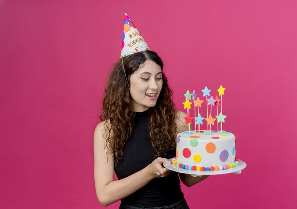美丽一个年轻漂亮的女人 卷发 戴着节日帽 手里拿着生日蛋糕 生日快乐 积极的生日派对概念 粉色的派对快乐蛋糕