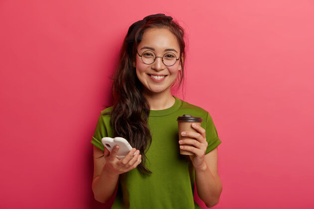 信息正面可爱的民族女孩在智能手机中滚动照片 使用现代智能手机和喝外卖咖啡 感觉感动和高兴 戴着圆眼镜 使用购物网站喜悦亚洲人人