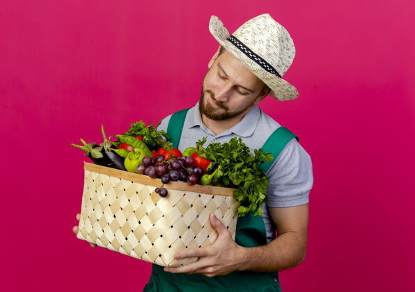年轻人高兴的年轻英俊的斯拉夫园丁穿着制服 戴着帽子 看着篮子里的蔬菜男人深红色斯拉夫人