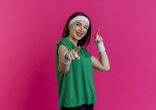 年轻快乐的年轻运动女性戴着头带和腕带站在侧视图眨眼看着做你的手势视图欢乐佩戴