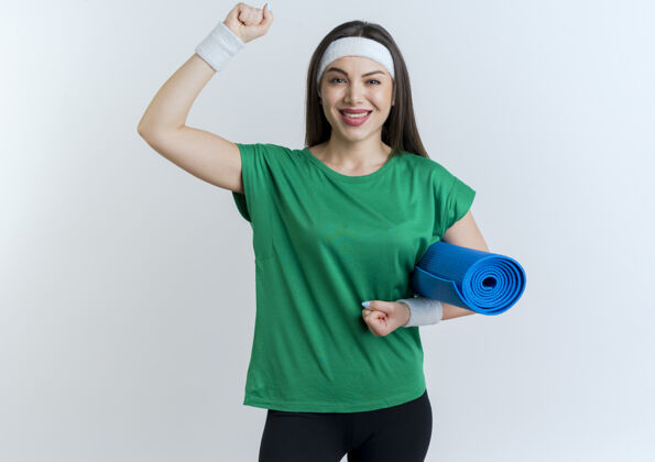 微笑微笑的年轻运动女性戴着头带和腕带看着拿着瑜伽垫举起拳头腕带向上女人